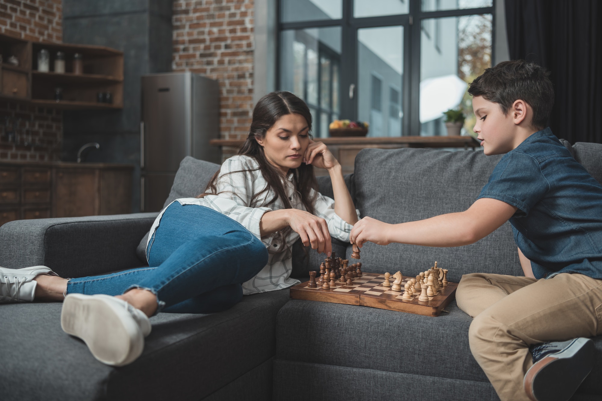 Little boy and his young mother playing chess on couch in living room